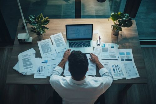 Osoba sedící u stolu plného papírů a otevřeného notebooku, obklopená rostlinami.